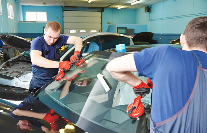Two technicians using suction tools to replace a car windshield in an auto repair garage.